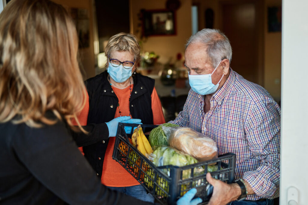 Senior couple on their 70s wearing a protective face mask picking up the groceries box that granddaughter is delivering in times of COVID-19, she is wearing mask and gloves.